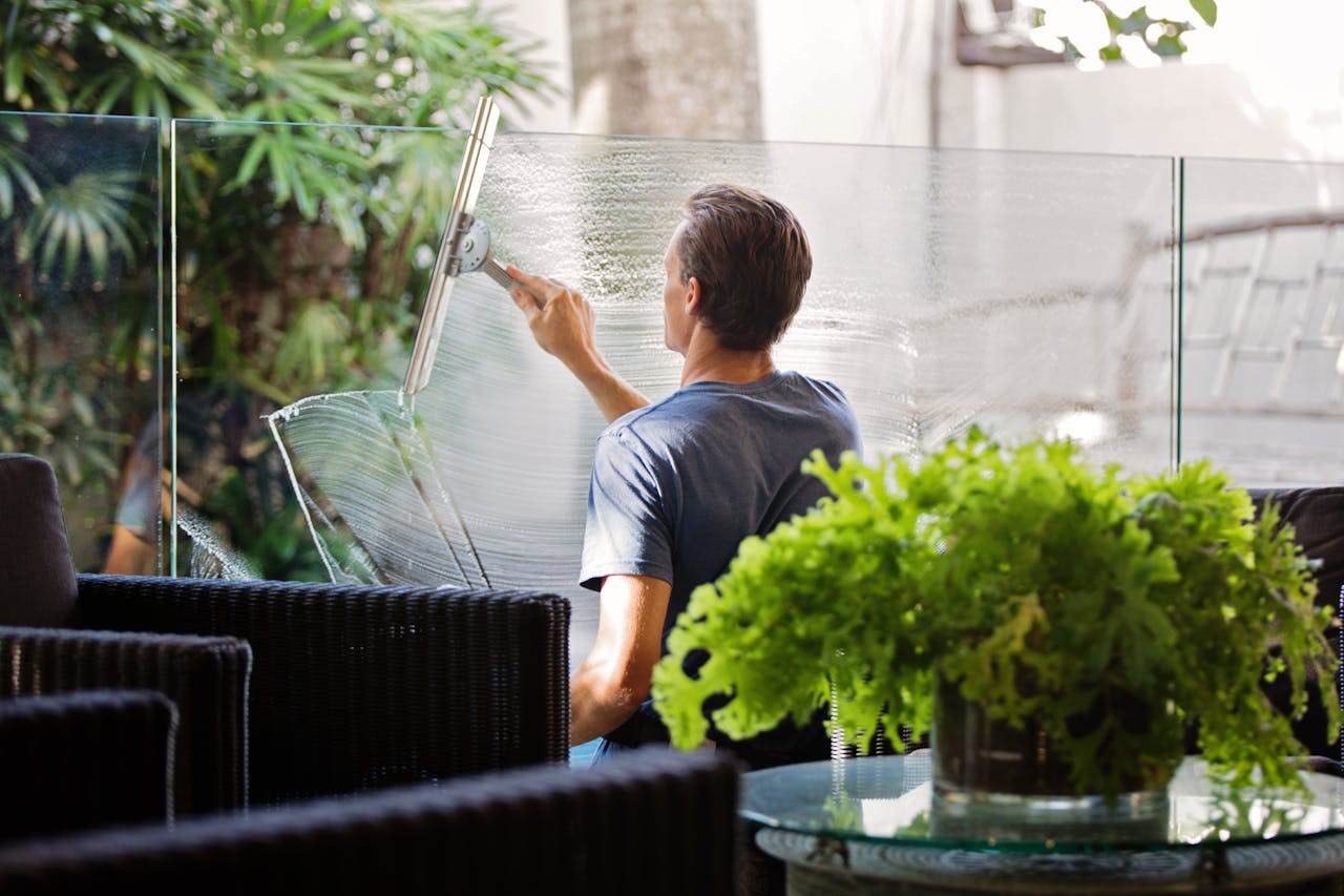 hero-img-01 A man cleaning a glass barrier outdoors with a squeegee, surrounded by greenery.
